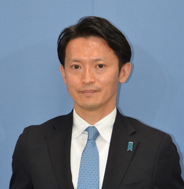 正面を向いて立つ日本人男性の顔写真です。ダークスーツと水色のネクタイを着用しており、髪は黒く整えられています。背景は単色の明るい色です。