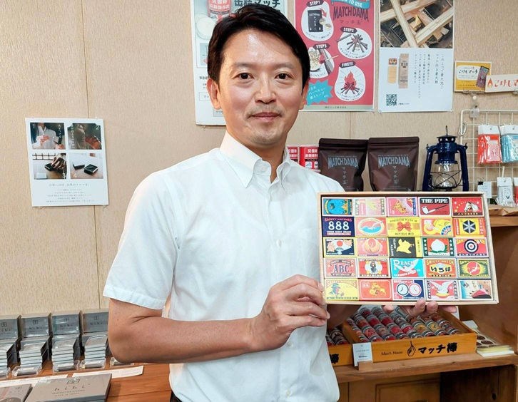 白い半袖シャツを着た日本人男性が、様々なデザインのパッケージが並んだ箱を持っている写真です。男性は軽く微笑んでおり、背景には商品が並べられた棚が見えます。