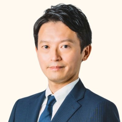 正面を向いて立つ日本人男性の顔写真です。ダークスーツと水色のネクタイを着用しており、髪は黒く整えられています。背景は単色の明るい色です。