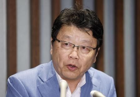 記者会見の場で、マイクが並んだテーブルの前に座る男性の姿です。青いジャケットを着ており、メガネをかけています。