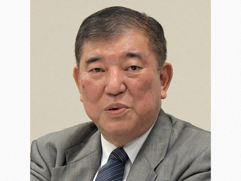 正面を向いて話している、グレーのスーツと青いネクタイを着用した中高年の男性のバストアップ写真です。

