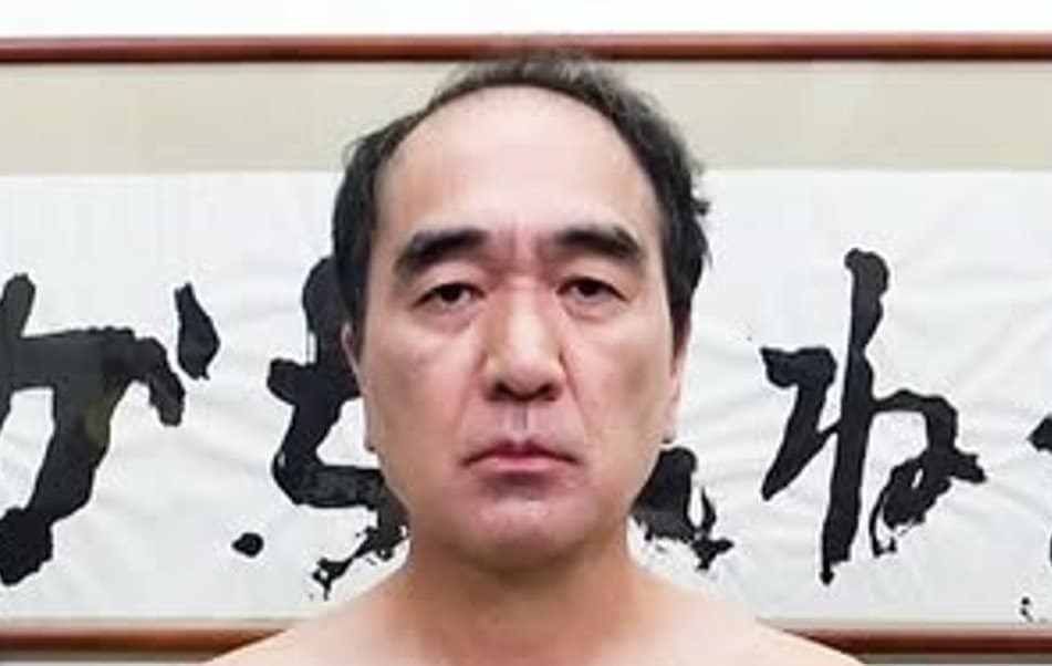 "背景に書道が飾られた場所で真顔でカメラを見つめている男性"
