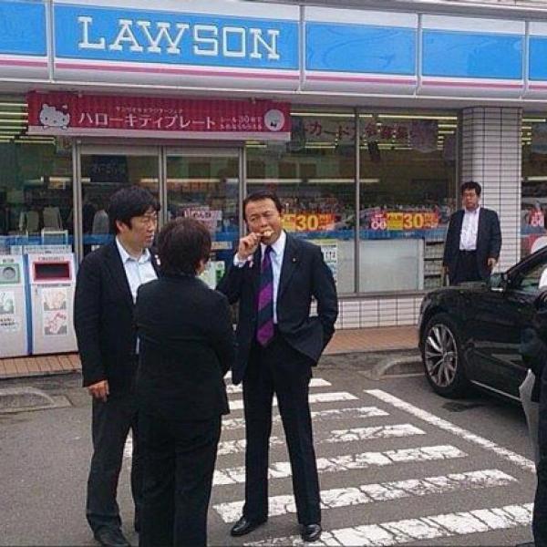 コンビニエンスストア「ローソン」の店先で、スーツ姿の男性政治家が棒アイスを食べている様子。周囲にはSPらしき人物がいる。