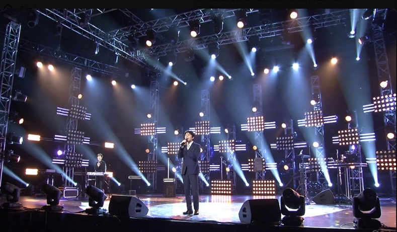 華やかな照明に包まれた音楽番組のスタジオセットで、司会者の男性がステージに立っている様子
